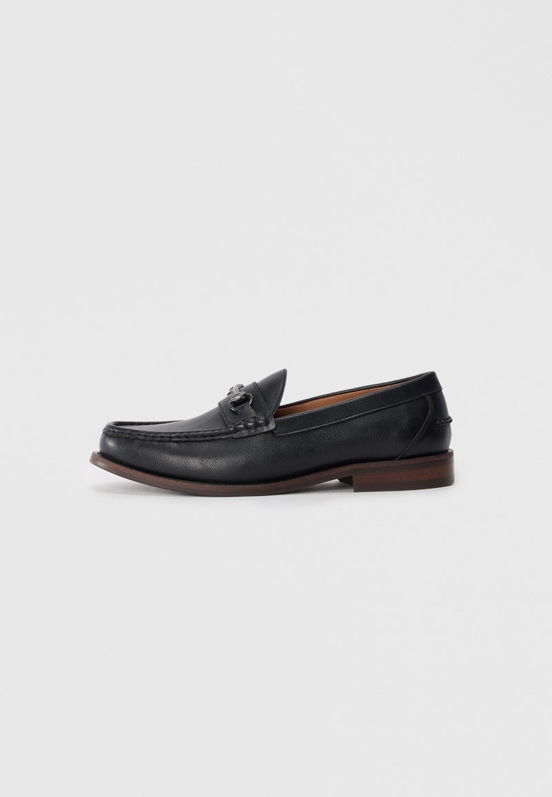Mocasines de cuero negro con punta redondeada, hardware decorativo en la parte delantera y una suela marrón contrastante. Textura suave y diseño fácil de poner.