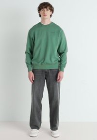 Groene sweatshirt met ronde halslijn, ribgebreide manchetten en een lichte textuur. Gecombineerd met loszittende grijze jeans en witte sneakers.