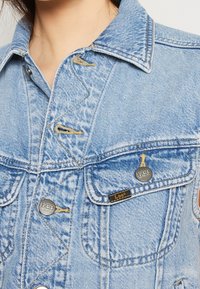 Jeansjacka i ljusblått med klassisk krage, metallknappar och bröstfickor. Har kontrasterande sömmar och strukturerad tyg.