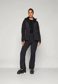 Casaco puffer preto com design acolchoado e detalhes em rosa, usado com calças pretas e botas de esqui. Apresenta capuz e bolsos com fecho.