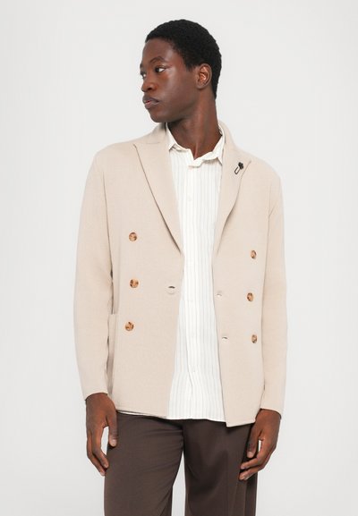 Beige Doppelreiherjacke mit Holzknöpfen, weichem Stoff und Reverskragen, getragen über einem weiß gestreiften Hemd und braunen Hosen.