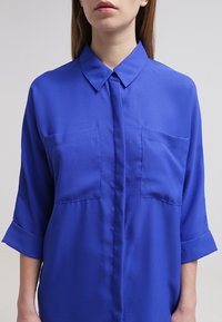 Blue, collared shirt in smooth fabric. Features two chest pockets, rolled sleeves, and a button-up front. Simple, classic design.