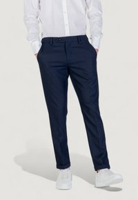 Personne portant un pantalon ajusté bleu marine, une chemise blanche à boutons avec les mains dans les poches, et des baskets blanches, se tenant contre un fond uni.