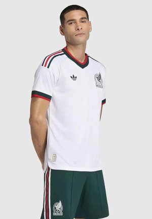 Mężczyzna w białej koszulce piłkarskiej Meksyku z zielonymi i czerwonymi akcentami oraz pasujące zielone spodenki z bocznymi paskami.