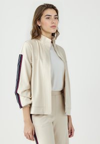 Beige Zip-jacke mit Stehkragen, die gestreifte Akzente in Dunkelblau und Burgunderrot an den Ärmeln aufweist, kombiniert mit passenden beige Hosen.
