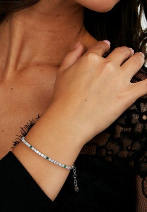 Silberarmband mit abwechselnd klaren und grünen Steinen, einer zarten Kettenakzent und einem Karabinerverschluss. Strukturierter schwarzer Spitzenhintergrund.