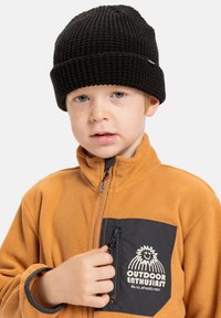 Černá pletená čepice na hlavě dítěte, doplněná béžovým fleecovým sakem s černou zipovou kapsou a grafickým logem.