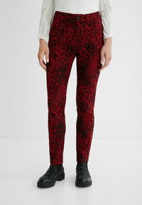 Vrouw gekleed in rode en zwarte panterprint broek met voorzakken, zwarte enkellaarzen en een witte gebreide top met lange mouwen.