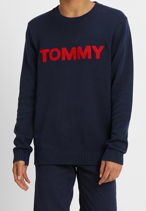 Pull bleu marine avec inscription rouge floue "TOMMY" sur la poitrine, porté avec un pantalon bleu marine assorti.