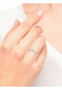 Anillo de banda plateada con diseño retorcido, que presenta pequeñas piedras preciosas transparentes, perfil texturizado, llevado en una mano con esmalte de uñas en tono neutro.