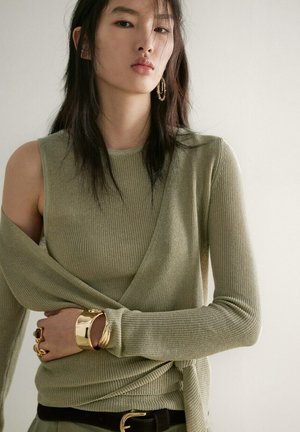 Jeune femme aux longs cheveux foncés porte un haut en maille vert olive avec des manches superposées, des boucles d’oreilles créoles dorées, un large bracelet et des bagues, posant contre un mur.