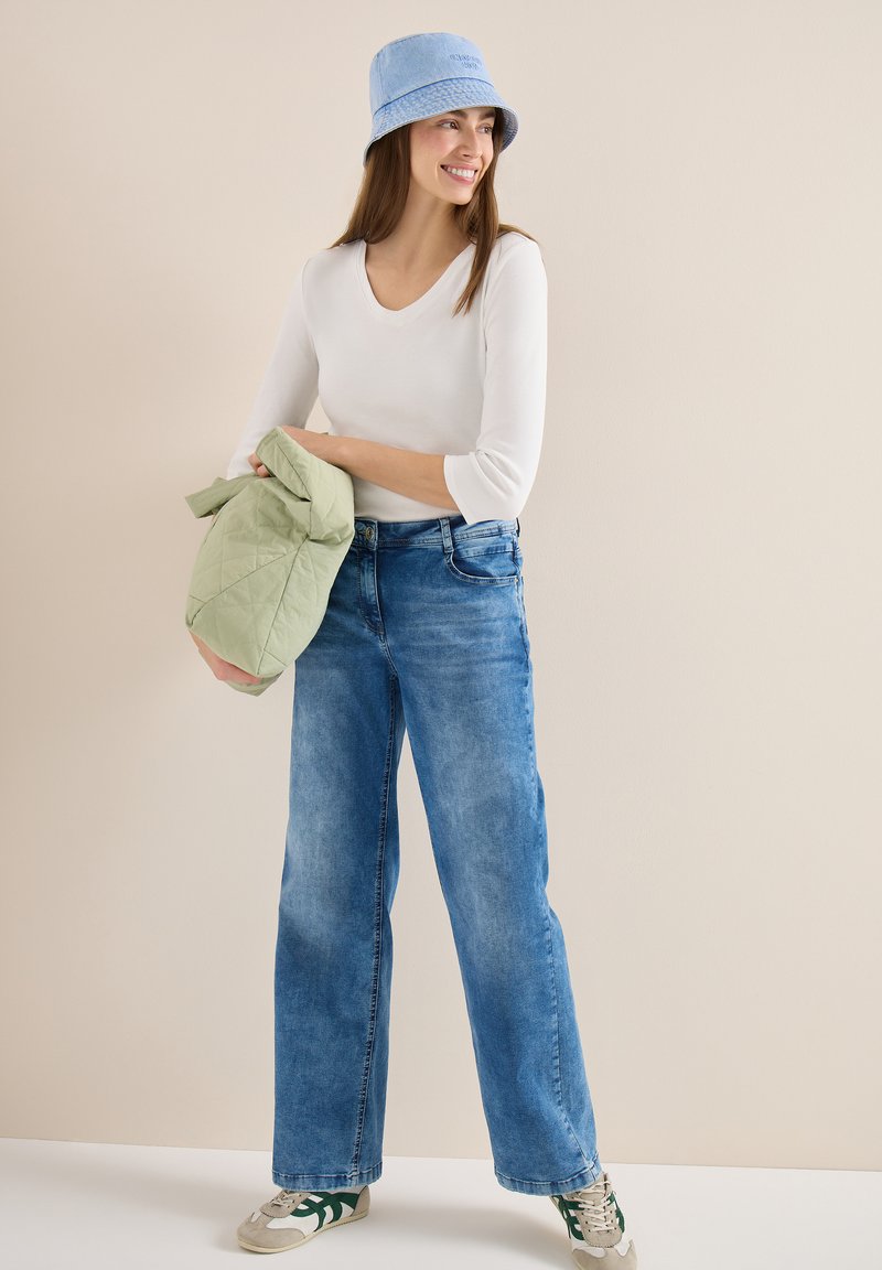 Femme souriante portant un chapeau seau bleu, une chemise blanche à manches longues, un jean à jambes larges et des baskets, tenant une veste matelassée vert clair.