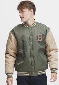 Giacca bomber verde oliva e beige con colletto a coste, chiusura frontale con bottoni a pressione e logo ricamato "B" sul petto. Texture morbida e imbottita.