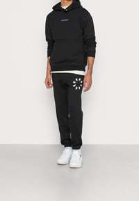 Man som bär en svart Adidas-hoodie med lila logotyp, matchande svarta sweatpants med vita trefoil-mönster och vita sneakers som står på en grå golv.