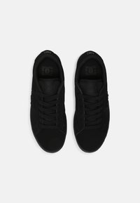 Zapatillas negras con una parte superior de tela suave, puntera redonda y cordones planos. Detalle sutil del logo en la lengüeta y el lado. Acentos texturizados en la suela.