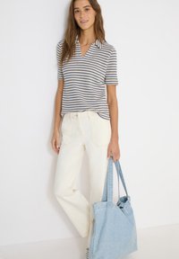 Femme portant une chemise à manches courtes rayée et un pantalon blanc, tenant un sac fourre-tout en denim bleu clair sur un fond blanc.