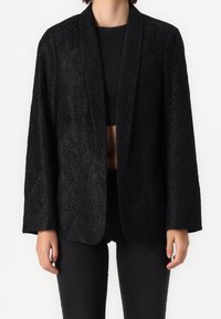 Femme portant un blazer noir texturé ouvert sur un crop top noir, associé à un pantalon noir, debout devant un fond uni.