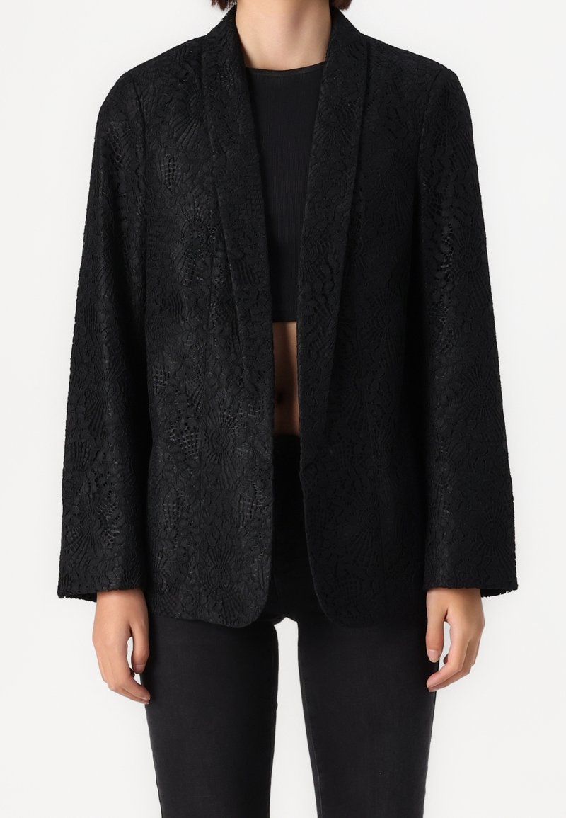 Femme portant un blazer noir texturé ouvert sur un crop top noir, associé à un pantalon noir, debout devant un fond uni.