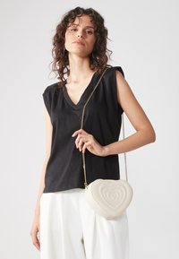 Czarny top bez rękawów z dekoltem w serek, zestawiony z kremową torebką w kształcie serca, ozdobioną przeszywanymi koncentrycznymi wzorami serc oraz złotym łańcuszkiem na ramię.