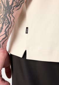 Kremfarget t-skjorte med sidedeling og svart "BOSS"-merke. Nærbilde av stofftekstur og tatovert arm. Brukt med mørke bukser.