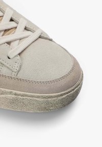 Zapatillas de ante beige claro con suela de goma gris texturizada, con detalles cosidos y cordones crema. Diseño de puntera redondeada.
