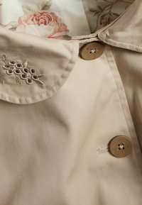 Beige trenchcoat med en kravehals, prydet med blomsterbroderi og en dekorativ brun knap. Glat tekstur, letvægtsstof.