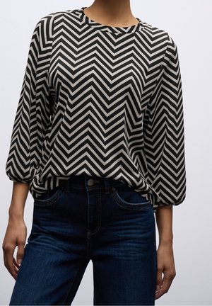 Kobieta ubrana w czarno-białą bluzkę z długim rękawem w zygzakowaty wzór, włożoną w ciemnogranatowe jeansy na gładkim tle.