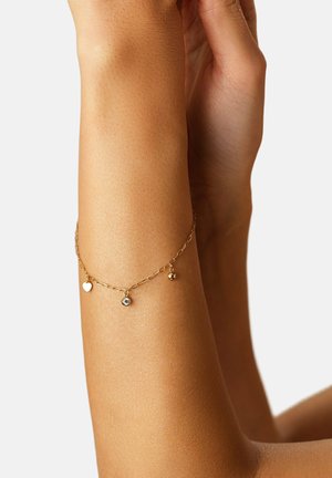 Handgelenk mit zartem Goldkettenarmband, das drei kleine Anhänger zeigt: Herz, klarer Edelstein und runde Perle, auf glatter Haut vor hellem Hintergrund.