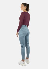 Bordeauxfarbener, langärmeliger Cropped-Top, kombiniert mit hellblauen, hochgeschnittenen Jeans. Weiße Sportschuhe runden das Outfit ab.