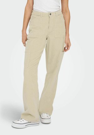 Beige Cord-Hosen mit lockerem Schnitt, ausgestattet mit zwei Fronttaschen und einem Knopfverschluss. Kombiniert mit weißen Sportschuhen.