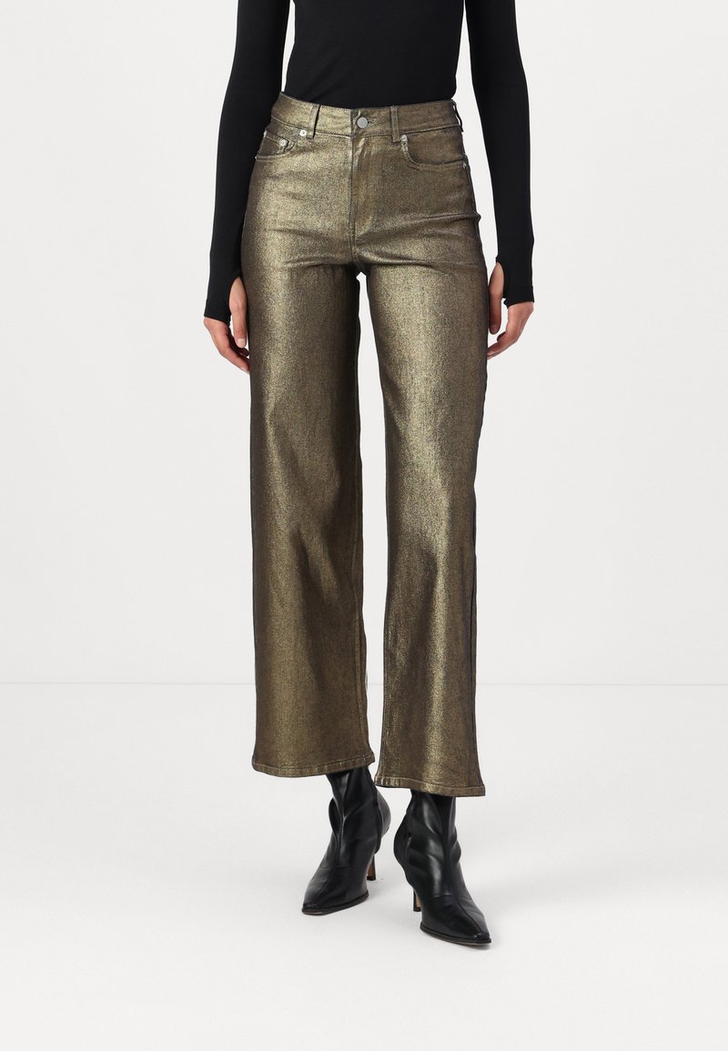Jeans anchos de metalizado dorado con cintura alta, con bolsillos frontales, cierre con cremallera y herrajes en plata. Combinados con botas de tobillo negras y de punta.