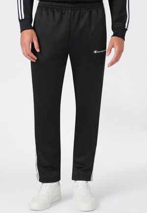 Pantaloni sportivi neri con strisce bianche laterali e logo Champion sulla parte superiore della coscia sinistra, indossati con scarpe da ginnastica bianche e top nero con strisce bianche.