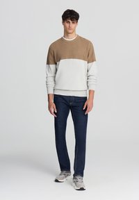 Sweter dzianinowy z brązową górną częścią i szaro-dolną, w połączeniu z ciemnoniebieskimi dżinsami i szarymi trampkami. Gładka tekstura, casualowy design.