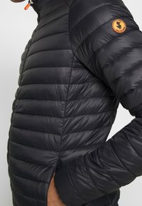 Giacca a vento nera con texture trapuntata, dotata di apertura con zip sul lato e un logo in gomma arancione sulla parte superiore del braccio.