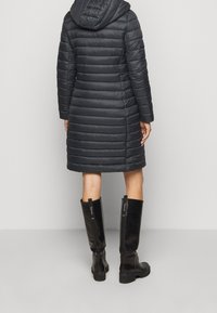 Sort dunjakke med hætte og synligt quiltet mønster, knælængde. Kombineret med knæhøje sorte glansfulde støvler med teksturerede detaljer.