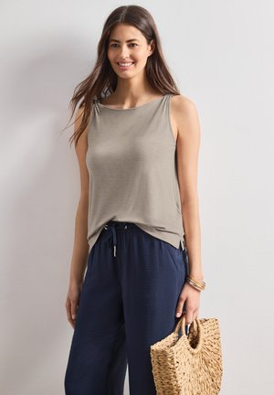 Lachende vrouw die een mouwloos beige topje en marineblauwe broek met trekkoord draagt, een gevlochten strohandtas vasthoudt en armbanden draagt.