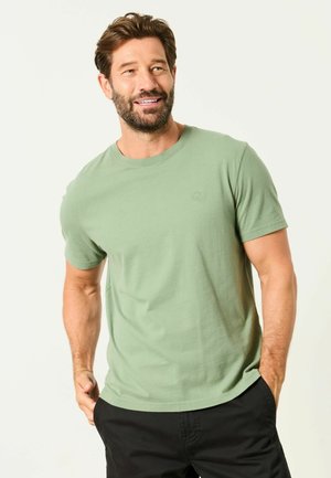 Smiling man with beard wearing light green short-sleeve shirt and black pants with hands in pockets against plain background.