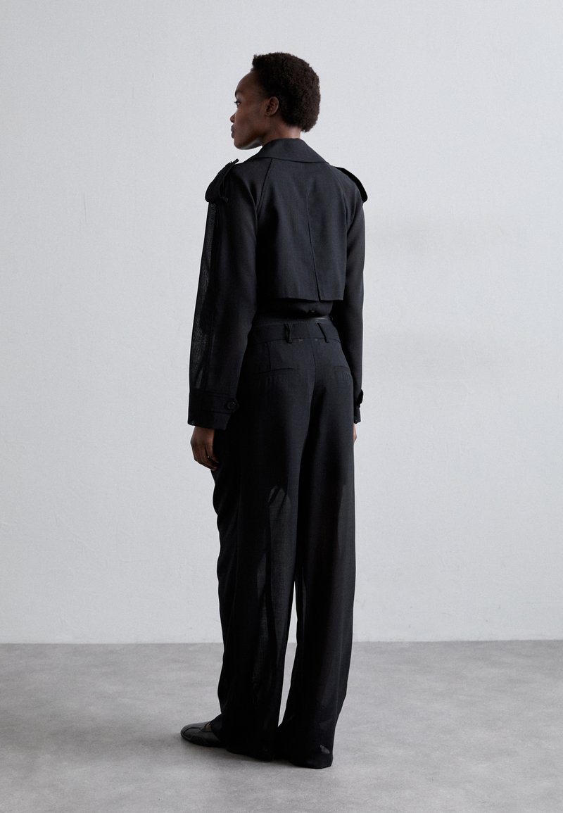 Black cropped jacket with structured shoulders and sheer sleeves. Paired with high-waisted black trousers. Smooth fabric texture.