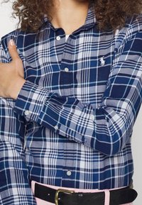 Kvinna som bär en blå och vit rutiga knäppskjorta med broderad logotyp, rosa byxor, svart bälte och armarna korsade över bröstet.