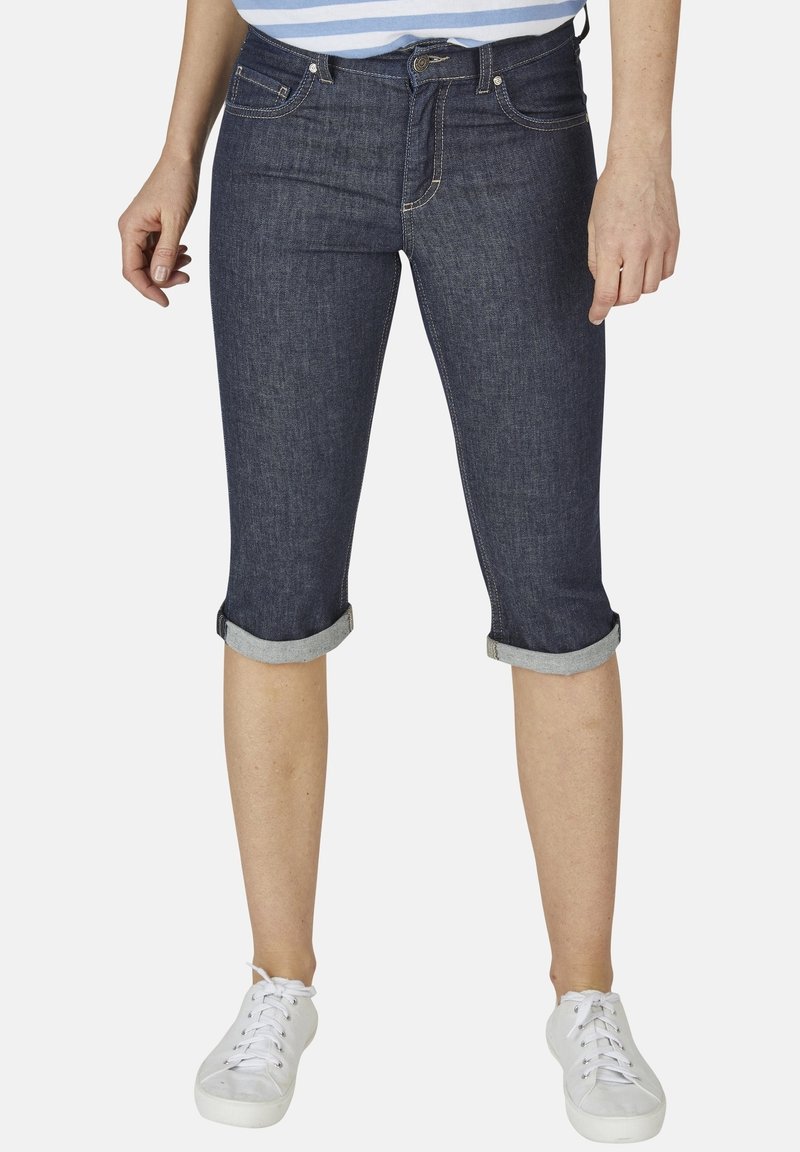 Femme portant un pantalon capri en denim bleu foncé retroussé et des baskets blanches sur un fond uni.