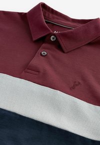 Burgundin värinen polo-paita, jossa on teksturoitu vaaleanharmaa raita ja laivastonsininen alaosa. Kolme nappia sisältävä napitus ja brodeerattu logo-detalji.