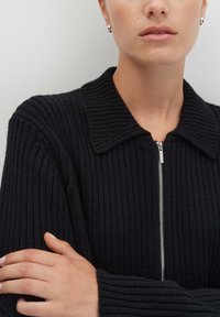 Vrouw in een zwarte ribgebreide zip-up trui met kraag, zilveren oorbellen, met de armen over elkaar tegen een effen lichtgrijze achtergrond.
