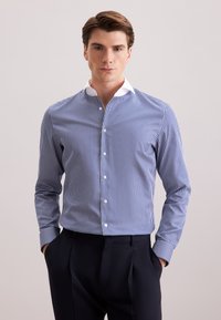 Uomo che indossa una camicia a righe verticali blu e bianche con colletto bianco e pantaloni neri, in piedi con le mani in tasca.