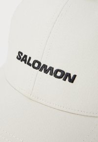 Kaki baseball cap laget av teksturert stoff, med svart brodert "SALOMON"-logo på forsiden, rund skjerm og sydd detaljer.
