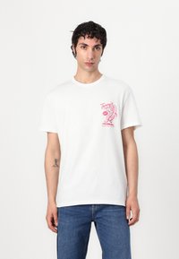 White cotton t-shirt with a crew neck, featuring a pink graphic print on the left chest area, paired with blue denim jeans.