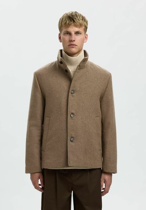 Giacca in misto lana marrone con colletto alto, pattina frontale, tre bottoni e tasche laterali, indossata sopra un maglione a maglia beige.