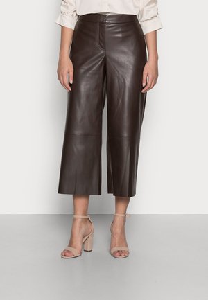 Vrouw draagt donkerbruine cropped wijde leren broek en beige hoge hak sandalen met enkelriem, staand tegen een witte muur.