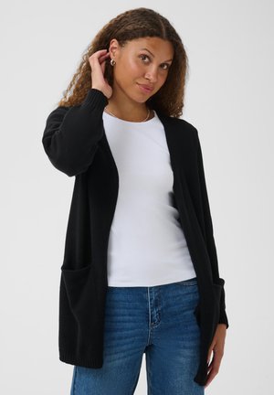 Jeune femme aux cheveux bouclés portant un cardigan noir, un t-shirt blanc et un jean bleu, touchant son oreille avec une main.