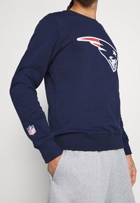 Sweatshirt bleu marine avec un grand logo d'équipe blanc et rouge sur le devant et un patch de la NFL sur la manche. Présente des poignets et un ourlet côtelés.
