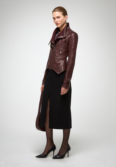 Veste de moto en cuir bordeaux au design ajusté, avec un ourlet asymétrique et des poches zippées, associée à une longue jupe noire et des escarpins noirs.